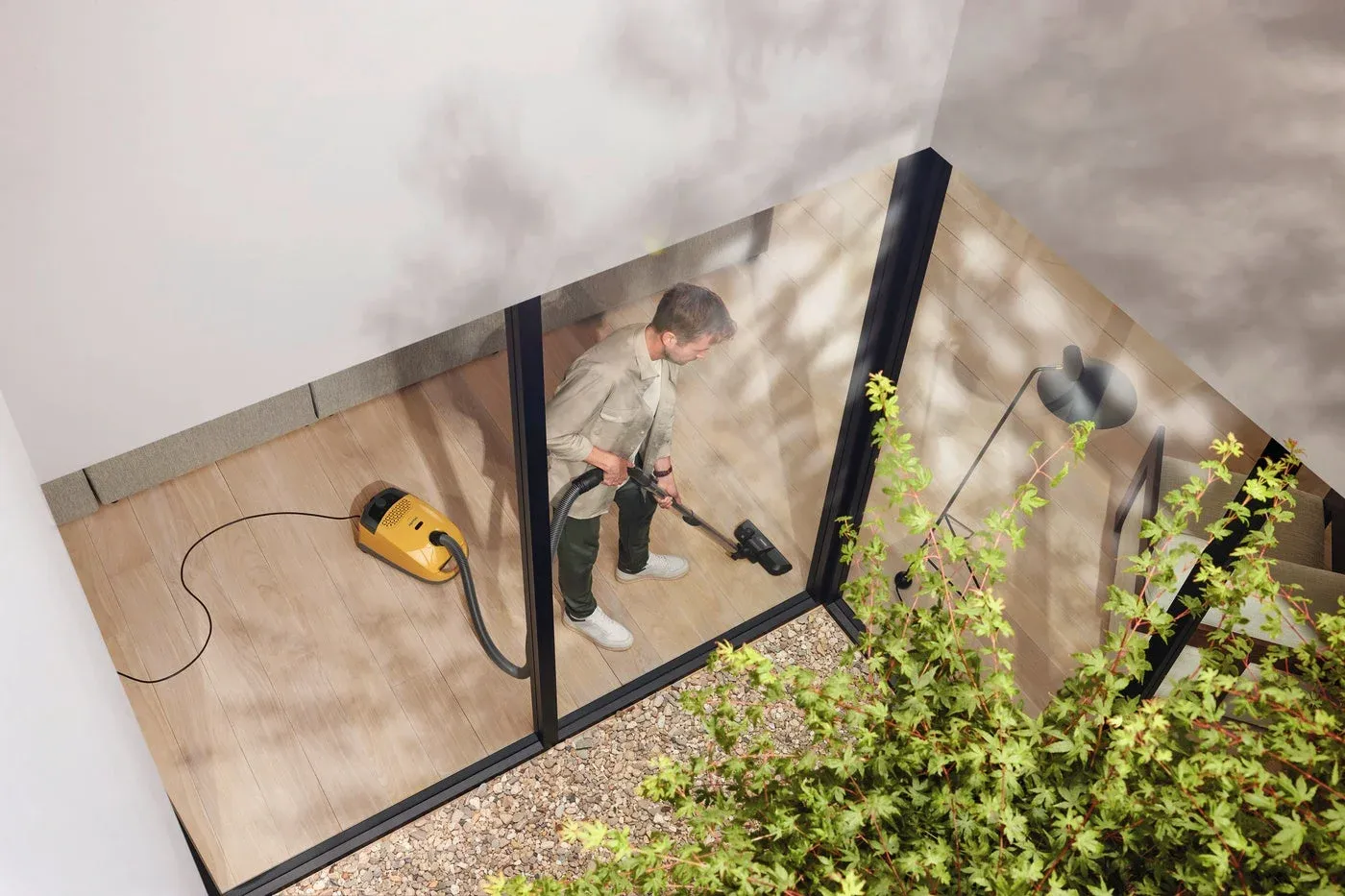 上から見た視点で、男性が黄色い掃除機を使ってフローリングを掃除している様子。大きな窓の外には植物と砂利が見える、明るくモダンな室内空間です。