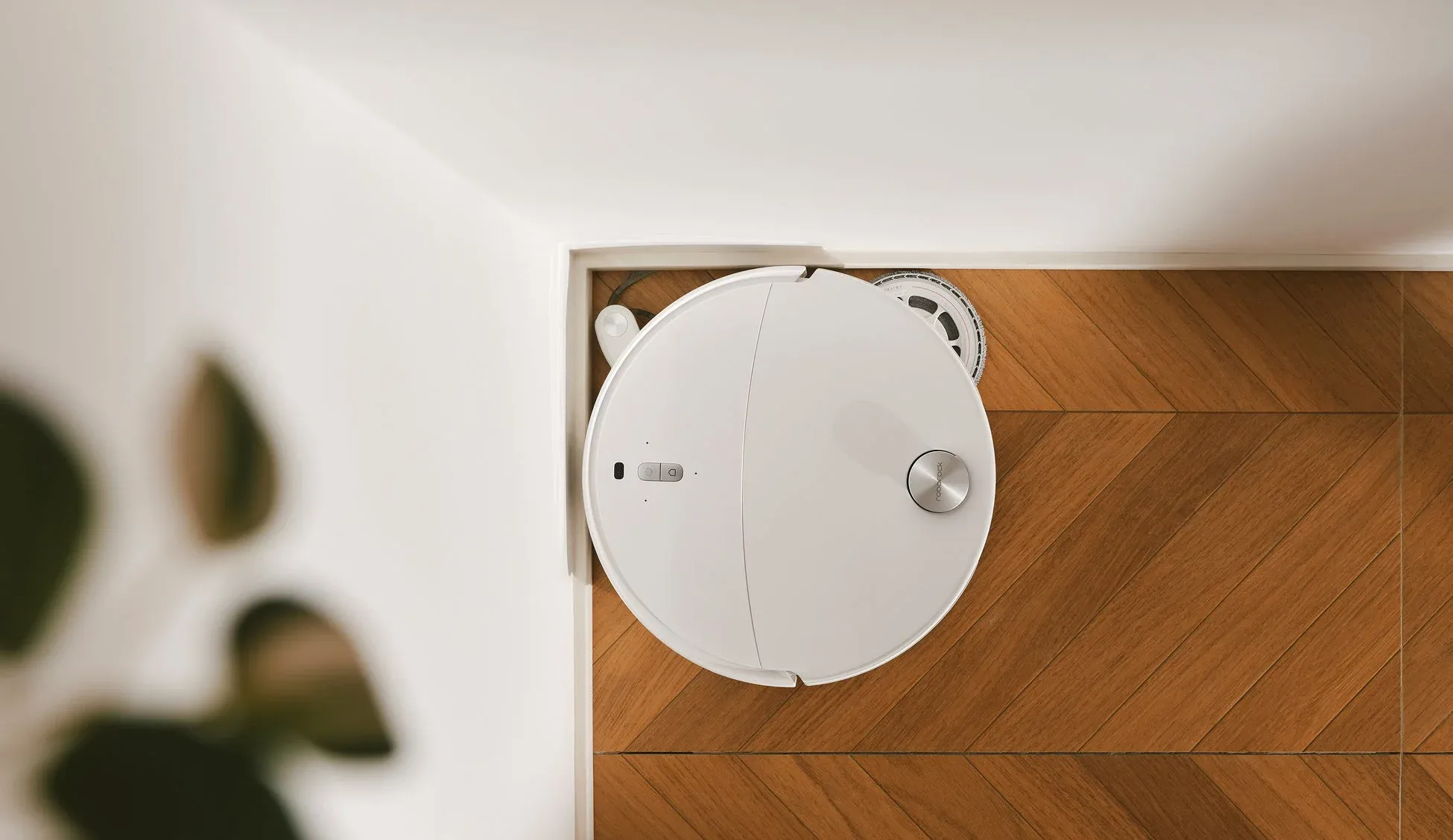 白い丸型のロボット掃除機が、ヘリンボーン柄の木製フローリングの上を掃除している様子を上から捉えた画像です。壁際に設置されており、スマートな室内風景を表現しています。