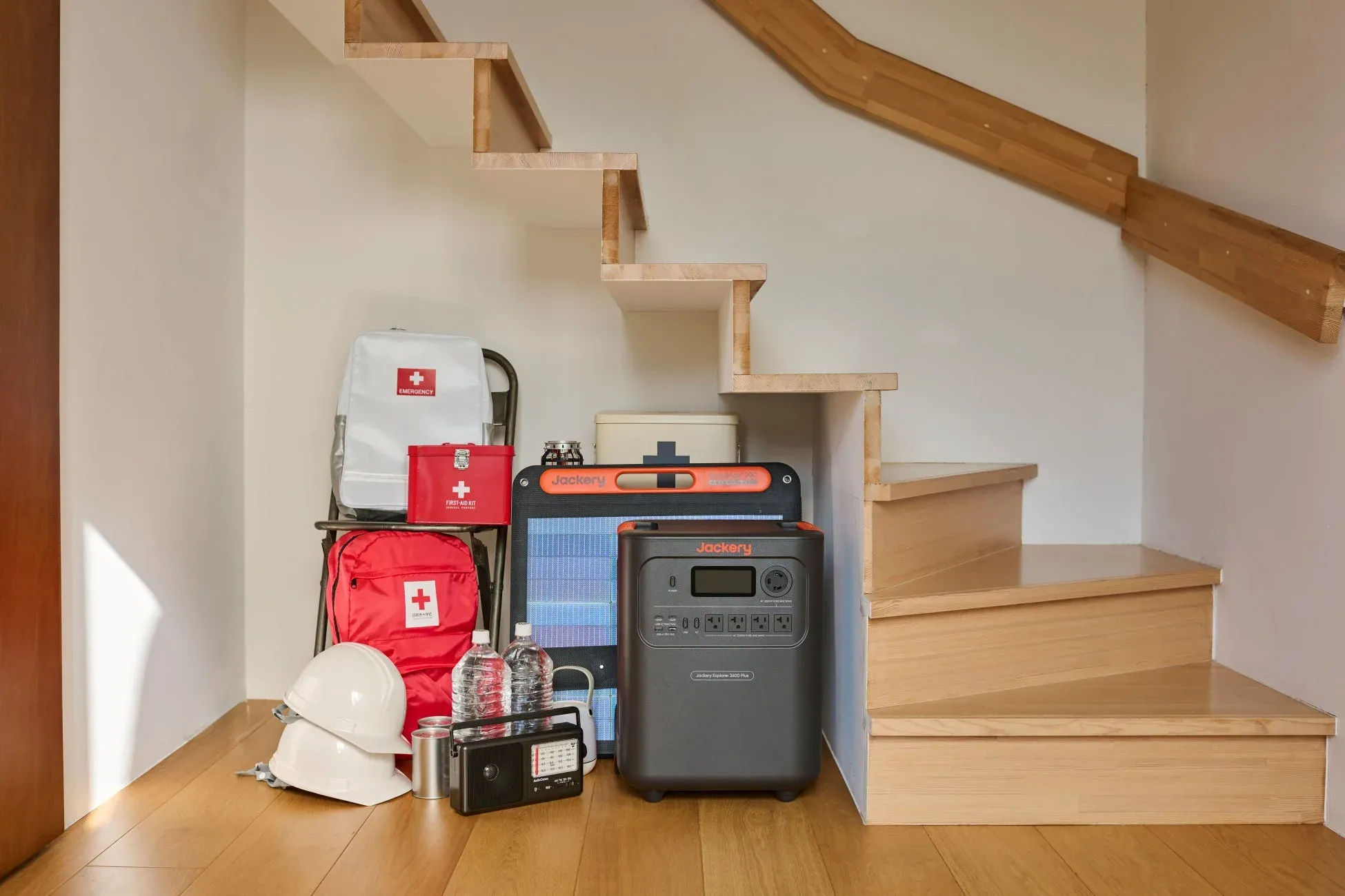 階段下にJackeryのポータブル電源とソーラーパネル、救急箱、ヘルメット、水、ラジオなどの防災用品が整然と置かれている