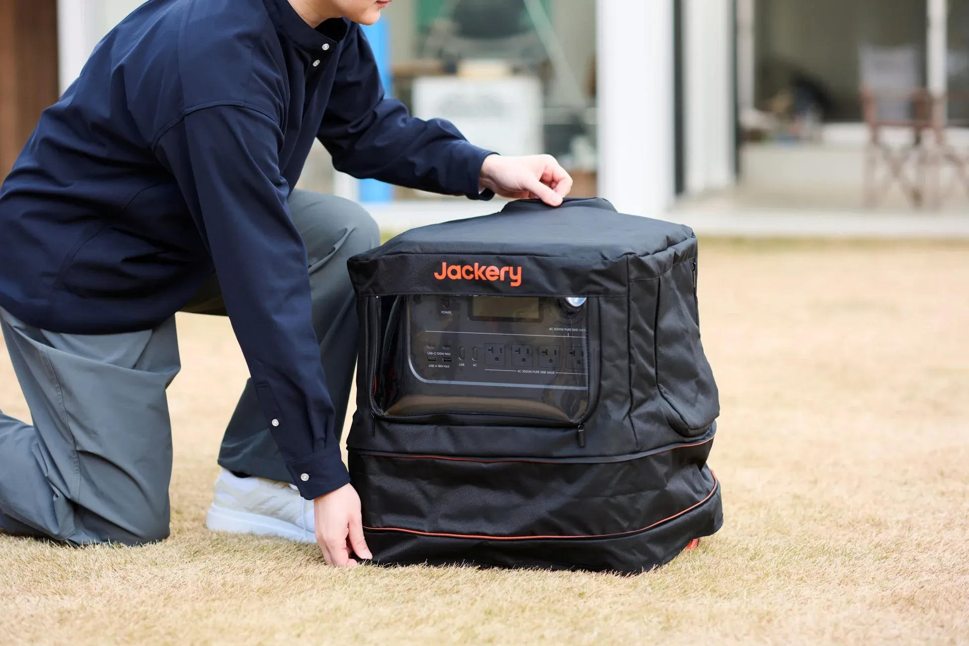 草地にしゃがんだ男性がJackeryのポータブル電源を操作しています。黒い保護ケースに入った電源のディスプレイやポートが見えています