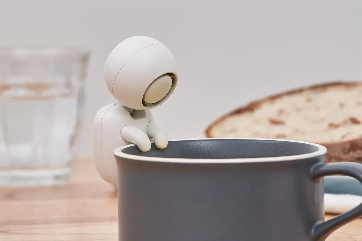 白いロボットのようなキャラクターが、ダークグレーのマグカップの縁にぶら下がり、中を覗き込んでいる様子