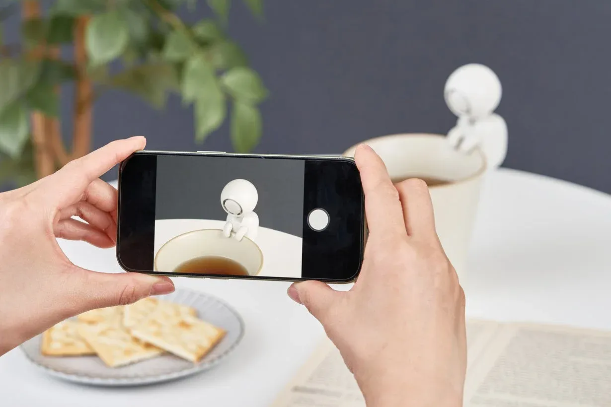 スマートフォンでティーカップの縁に座る白いミニチュアフィギュアを撮影している様子