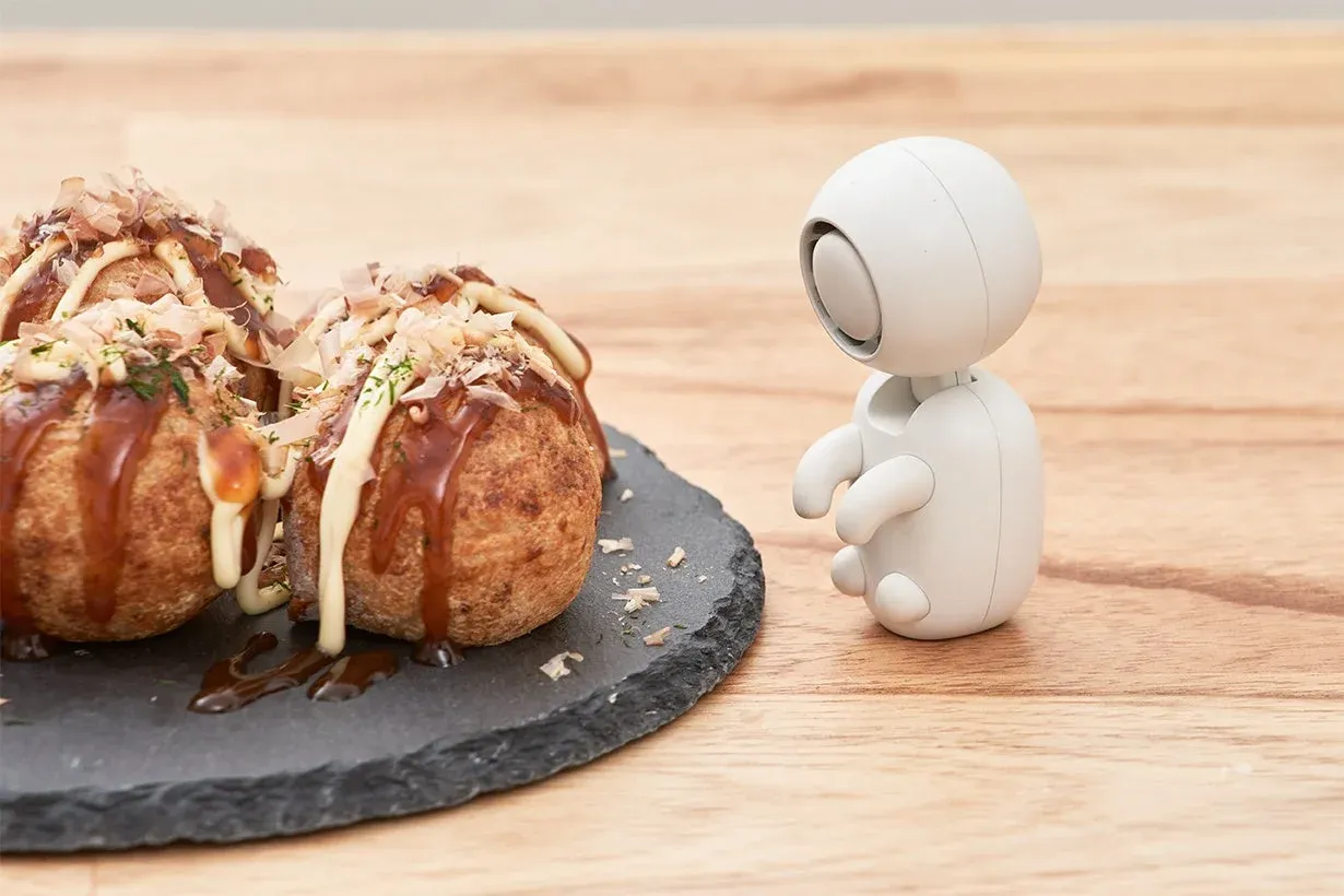 美味しそうな熱々のたこ焼きが黒いプレートに盛られ、その横で白い小型ロボットがじっと見つめている