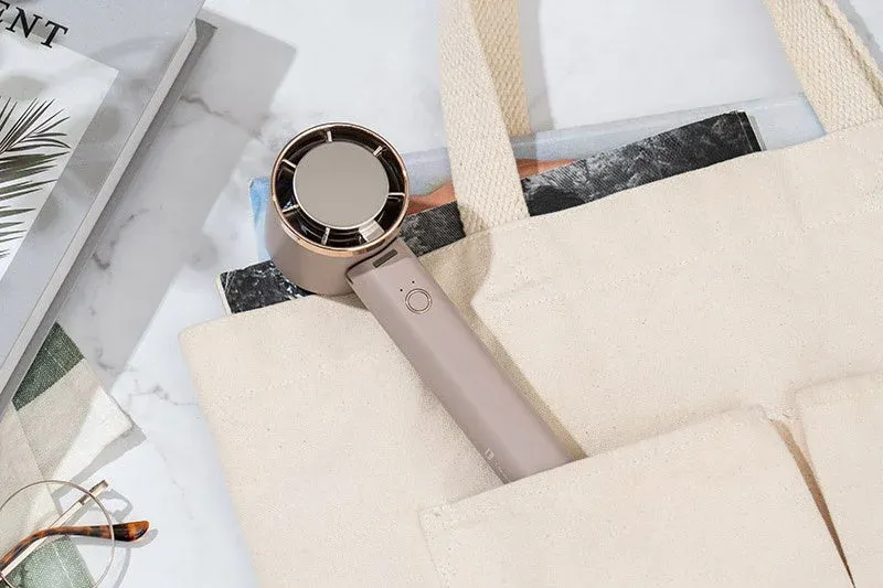 ベージュ色のハンディファンがキャンバス地のトートバッグのポケットから顔を出し、その周りには本やメガネが置かれた大理石のテーブルの上で、日常の一コマを切り取ったようなおしゃれな雰囲気の写真