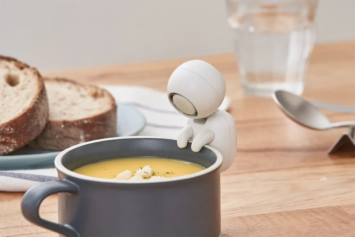 マグカップに入った温かいスープを、可愛らしい白いロボットが覗き込んでいる様子