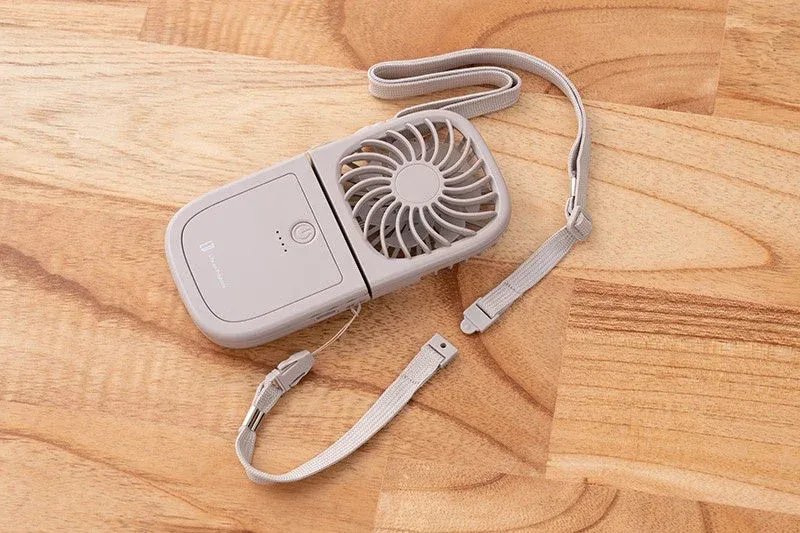 グレーの折りたたみ式携帯扇風機が木製のテーブルに置かれています。電源ボタンとバッテリー残量を示すLED、そして持ち運び用のストラップが見えます。夏の外出時に便利なアイテムです。