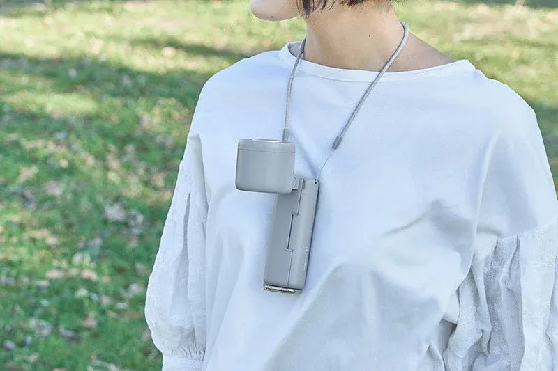 白いトップスを着た人物が、屋外でグレーの首掛け扇風機を首に下げて使用している様子