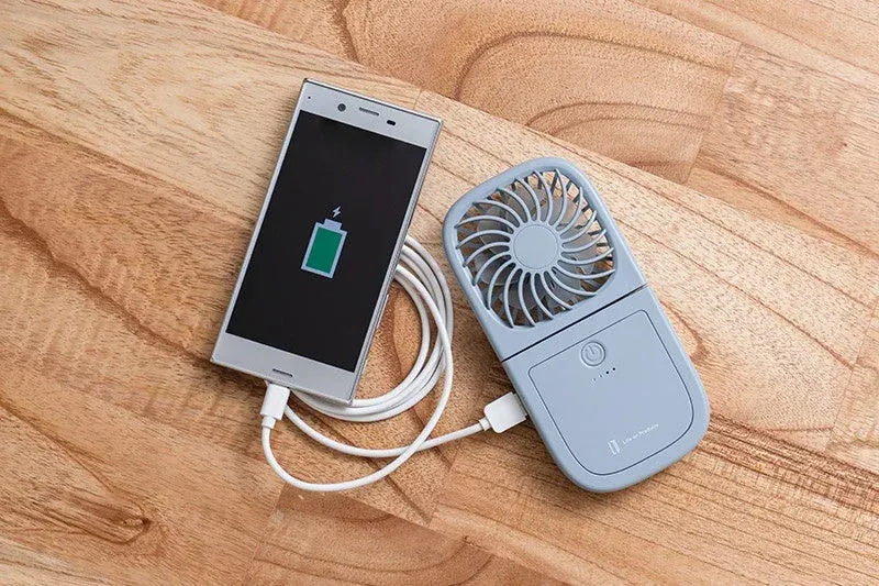 木製の床に置かれたスマートフォンが、ポータブル扇風機兼モバイルバッテリーからUSBケーブルで充電されている様子。外出先での充電と冷却を両立する便利なガジェットの組み合わせを示しています。