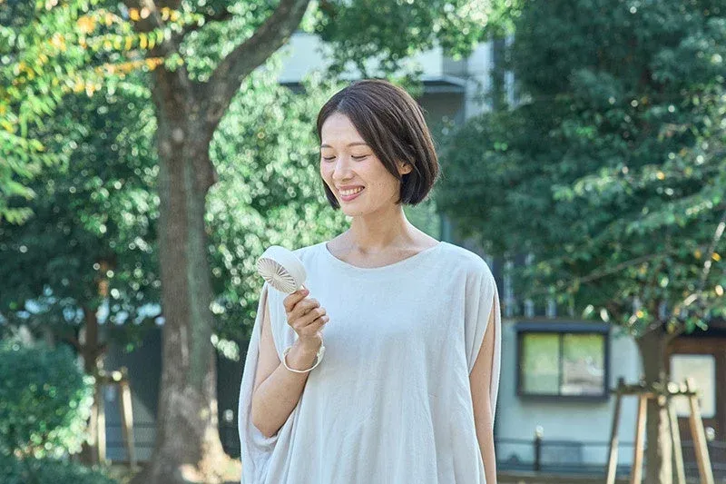 緑豊かな屋外で、笑顔の女性が白いハンディファンを持ち、涼んでいる様子です。