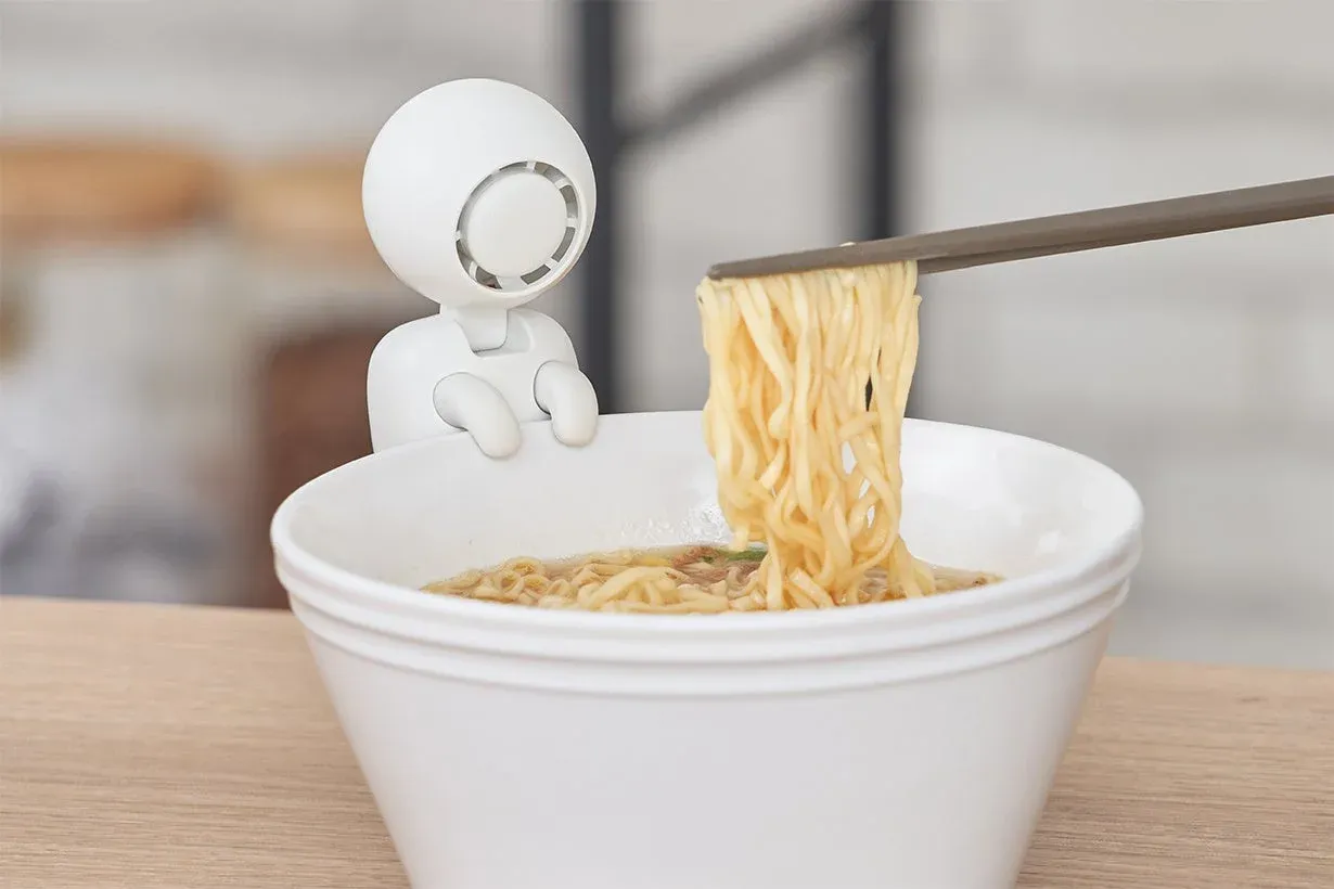 白い器に入ったインスタントラーメンを箸で持ち上げている様子です。器の縁には、白い可愛らしい人型キャラクターのガジェットが座って見守っています。