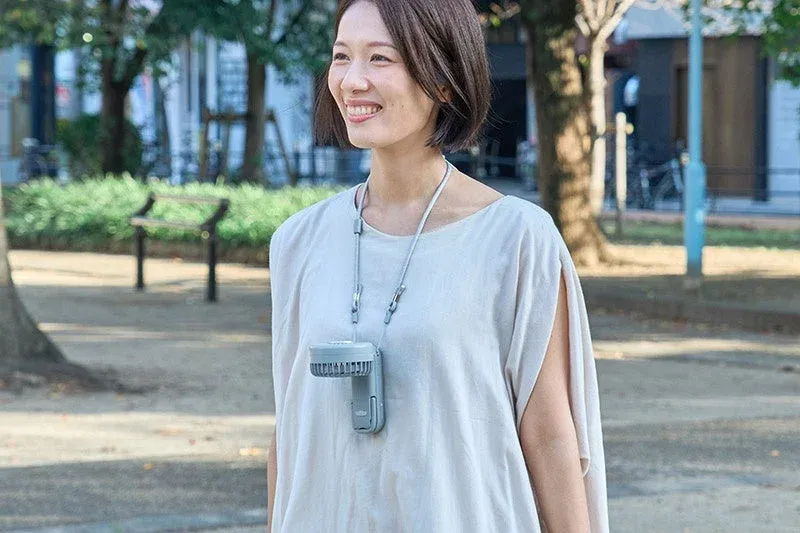 屋外で、笑顔の女性が首から携帯型のネックファンを下げている